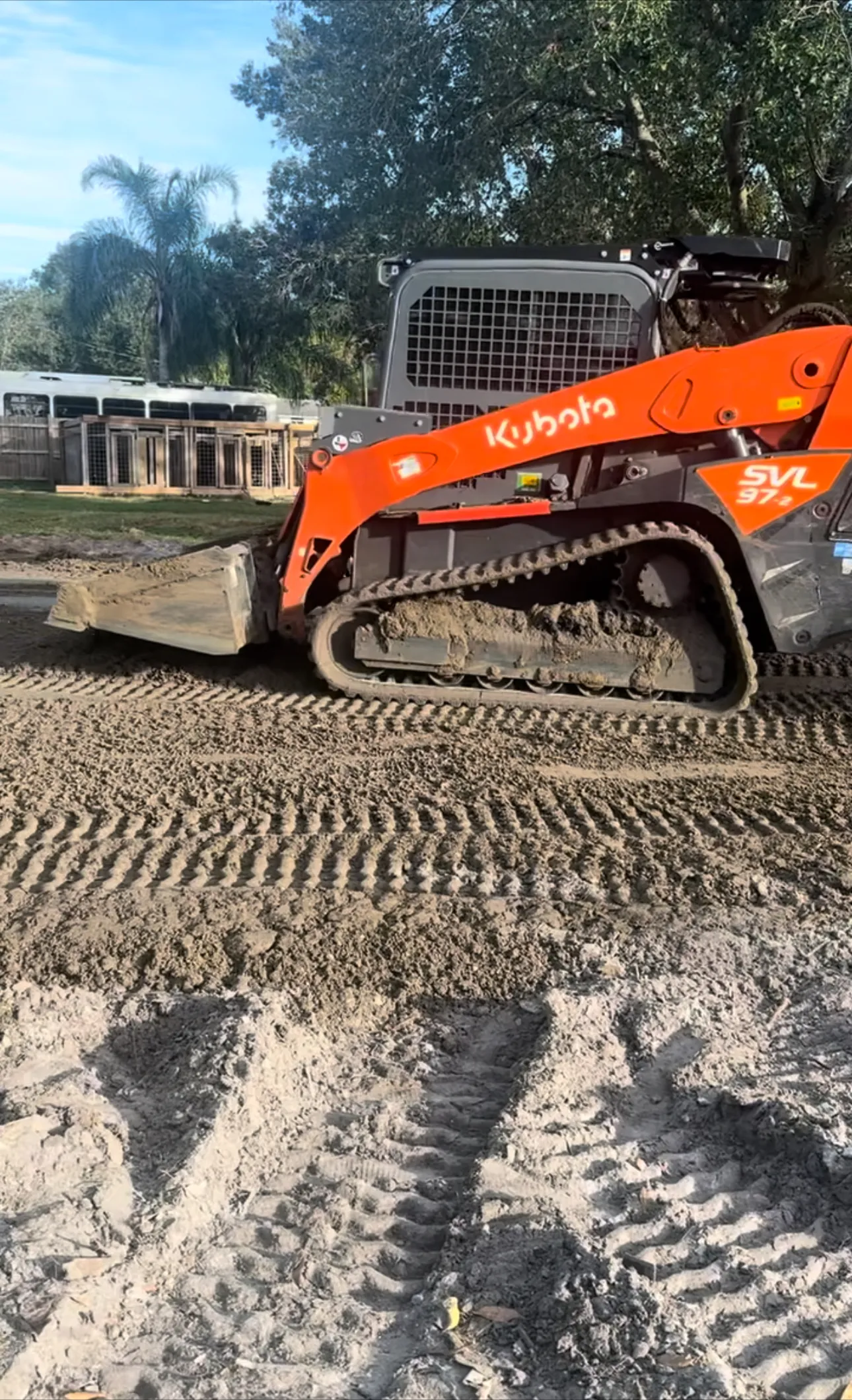 Orange compact track loader leveling sandy soil on a lot, with trees and a building behind.