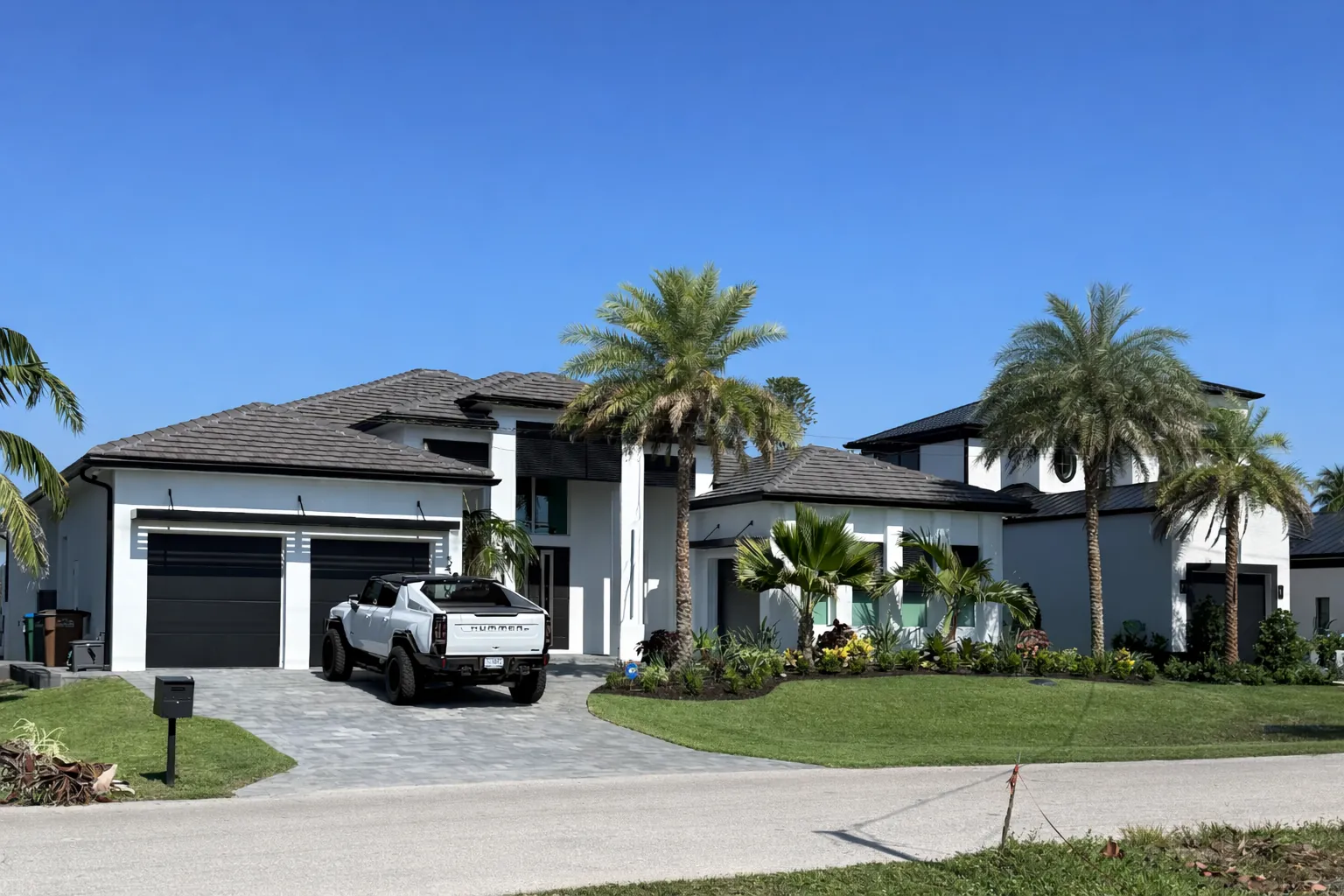 Modern white home with a dark tile roof, black garage doors, paver driveway, palm trees and a parked truck.